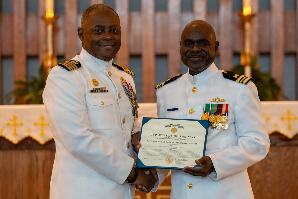 Two U.S. Navy officers in white dress uniforms pose together holding a Department of the Navy certificate. The certificate visible in the image is a Navy and Marine Corps Commendation Medal award. The ceremony appears to be taking place in a chapel or religious setting based on the altar and crosses visible in the background.