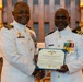 Two U.S. Navy officers in white dress uniforms pose together holding a Department of the Navy certificate. The certificate visible in the image is a Navy and Marine Corps Commendation Medal award. The ceremony appears to be taking place in a chapel or religious setting based on the altar and crosses visible in the background.