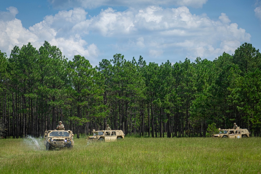 A convoy of three tan/desert-camouflage military vehicles, including what appear to be HMMWVs (Humvees) and at least one JLTV or similar tactical vehicle, traverse an open grassy field. Personnel are visible manning turret positions atop the vehicles. The setting appears to be a training area with a dense pine forest backdrop and partly cloudy sky.