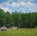 A convoy of three tan/desert-camouflage military vehicles, including what appear to be HMMWVs (Humvees) and at least one JLTV or similar tactical vehicle, traverse an open grassy field. Personnel are visible manning turret positions atop the vehicles. The setting appears to be a training area with a dense pine forest backdrop and partly cloudy sky.