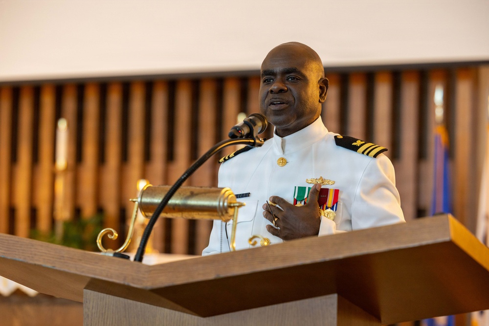 A naval officer in white dress uniform is speaking at a podium equipped with a microphone. He is wearing multiple service ribbons, gold insignia, and shoulder boards indicating a senior rank, likely a Navy Captain (O-6). The setting appears to be a formal ceremony, possibly in a chapel or ceremonial hall.