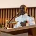 A naval officer in white dress uniform is speaking at a podium equipped with a microphone. He is wearing multiple service ribbons, gold insignia, and shoulder boards indicating a senior rank, likely a Navy Captain (O-6). The setting appears to be a formal ceremony, possibly in a chapel or ceremonial hall.