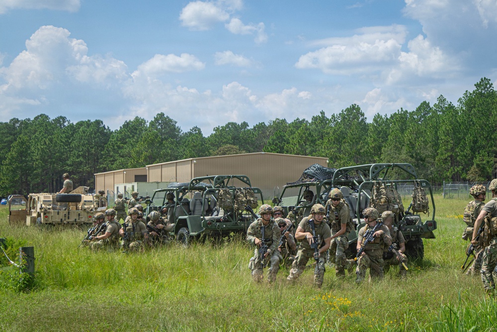 A group of military personnel in camouflage uniforms are assembled in an open grassy field alongside multiple military utility vehicles, including what appears to be Polaris MRZR-type all-terrain vehicles and a HMMWV (Humvee). The soldiers are armed and appear to be staging or preparing for a training exercise. The setting includes a tan warehouse-style building and a treeline of pine trees in the background, consistent with a military training installation.