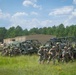 A group of military personnel in camouflage uniforms are assembled in an open grassy field alongside multiple military utility vehicles, including what appears to be Polaris MRZR-type all-terrain vehicles and a HMMWV (Humvee). The soldiers are armed and appear to be staging or preparing for a training exercise. The setting includes a tan warehouse-style building and a treeline of pine trees in the background, consistent with a military training installation.