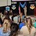 A military service member in OCP (Operational Camouflage Pattern) uniform is addressing a seated civilian audience at what appears to be a formal briefing or family readiness event. The setting is a military hangar or large facility decorated with Air Force unit insignia and flags. The visible emblems identify this as a 445th Airlift Wing and Air Force Reserve Command event.