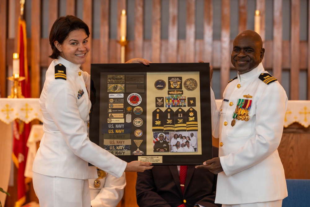 Two U.S. Navy officers in white dress uniforms are holding a framed shadow box containing military memorabilia, patches, rank insignia, medals, and photographs. The ceremony appears to be a retirement or recognition event, likely taking place in a chapel or ceremonial hall. The shadow box commemorates the career of a Navy Supply Corps officer, with visible 'U.S. NAVY' name tapes and rank epaulettes displayed.