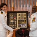 Two U.S. Navy officers in white dress uniforms are holding a framed shadow box containing military memorabilia, patches, rank insignia, medals, and photographs. The ceremony appears to be a retirement or recognition event, likely taking place in a chapel or ceremonial hall. The shadow box commemorates the career of a Navy Supply Corps officer, with visible 'U.S. NAVY' name tapes and rank epaulettes displayed.