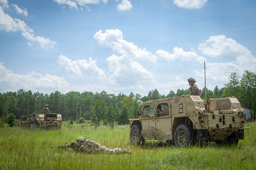 A military field training exercise is underway in an open grassy area bordered by pine trees. Two tan/desert-camouflage tactical vehicles, appearing to be Joint Light Tactical Vehicles (JLTVs), are positioned in the field with soldiers manning them and one soldier in a prone firing position on the ground. The scene suggests a live-fire or tactical movement exercise conducted in a temperate, forested training environment.