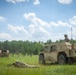 A military field training exercise is underway in an open grassy area bordered by pine trees. Two tan/desert-camouflage tactical vehicles, appearing to be Joint Light Tactical Vehicles (JLTVs), are positioned in the field with soldiers manning them and one soldier in a prone firing position on the ground. The scene suggests a live-fire or tactical movement exercise conducted in a temperate, forested training environment.