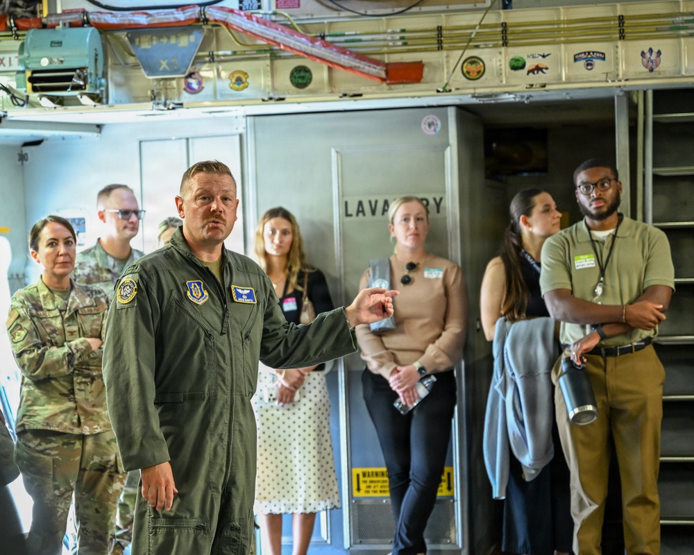 A U.S. Air Force officer in a green flight suit is briefing or giving a tour to a group of civilians and military personnel inside what appears to be a large military aircraft, likely a C-17 or similar transport aircraft. The officer is gesturing while speaking, with several attendees wearing name badge lanyards listening attentively. The interior shows aircraft fixtures including a lavatory compartment, overhead equipment, and unit stickers/patches on the ceiling panels.