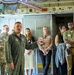 A U.S. Air Force officer in a green flight suit is briefing or giving a tour to a group of civilians and military personnel inside what appears to be a large military aircraft, likely a C-17 or similar transport aircraft. The officer is gesturing while speaking, with several attendees wearing name badge lanyards listening attentively. The interior shows aircraft fixtures including a lavatory compartment, overhead equipment, and unit stickers/patches on the ceiling panels.