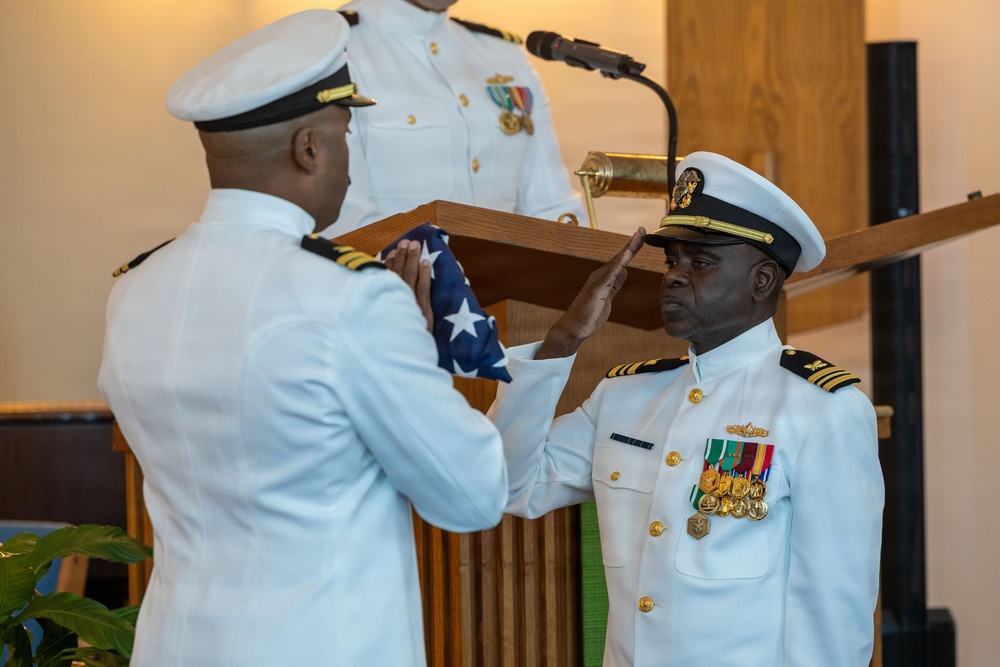 A formal U.S. Navy military ceremony is taking place, likely a retirement ceremony. One naval officer is presenting a folded American flag to another officer who is rendering a salute. A third uniformed individual stands at a podium with a microphone in the background.