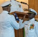 A formal U.S. Navy military ceremony is taking place, likely a retirement ceremony. One naval officer is presenting a folded American flag to another officer who is rendering a salute. A third uniformed individual stands at a podium with a microphone in the background.