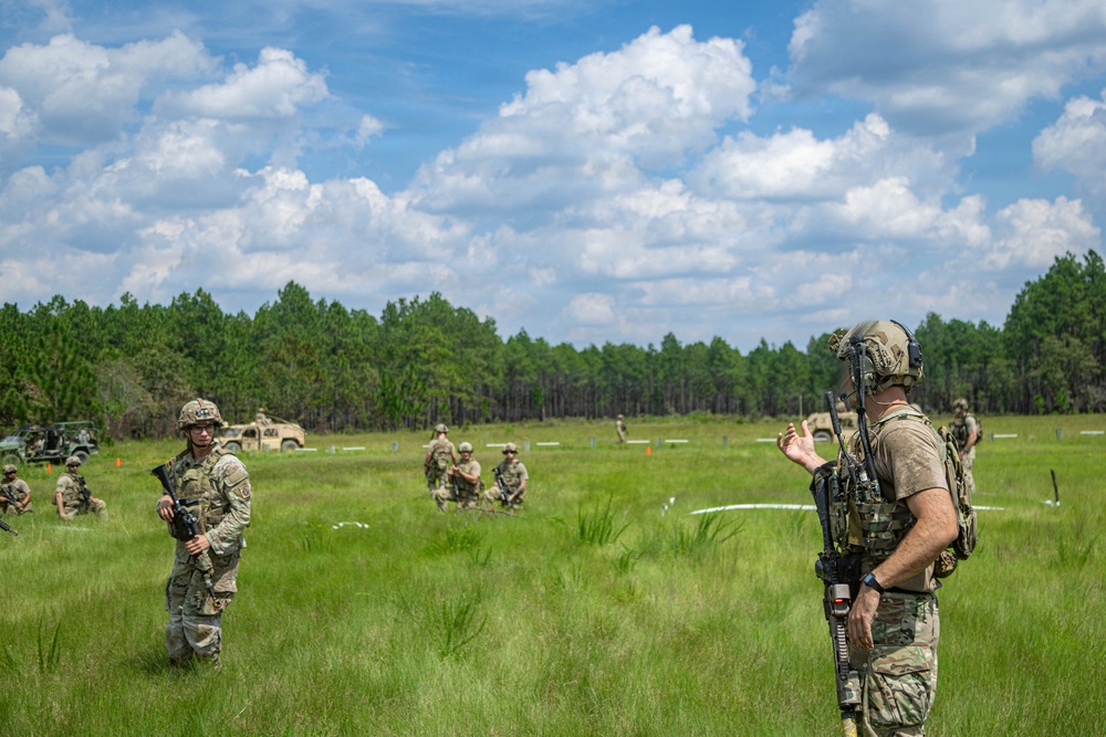 Military personnel in camouflage uniforms are conducting a field training exercise in an open grassy area. A soldier in the foreground appears to be instructing or briefing others who are spread across the field. Military vehicles are visible in the background near a tree line of tall pine trees.