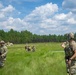 Military personnel in camouflage uniforms are conducting a field training exercise in an open grassy area. A soldier in the foreground appears to be instructing or briefing others who are spread across the field. Military vehicles are visible in the background near a tree line of tall pine trees.