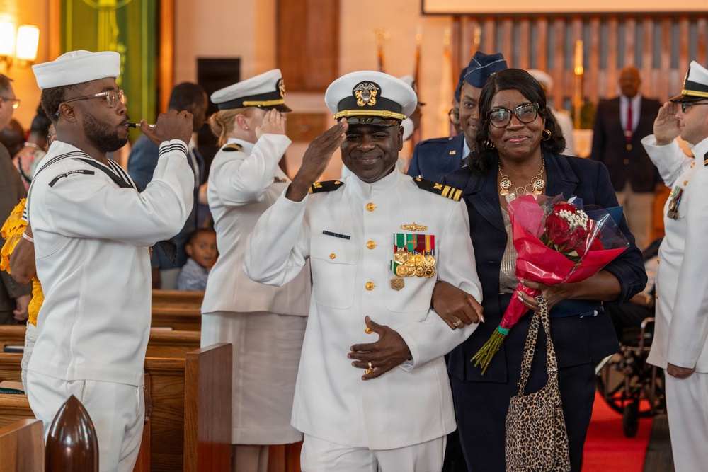 A military ceremony taking place inside what appears to be a church or chapel, featuring a senior Navy officer in dress whites being honored alongside a woman holding a bouquet of red flowers. Multiple service members in various uniforms are rendering salutes, suggesting a retirement ceremony or formal recognition event. The setting and attire indicate a formal, celebratory military occasion.