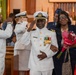 A military ceremony taking place inside what appears to be a church or chapel, featuring a senior Navy officer in dress whites being honored alongside a woman holding a bouquet of red flowers. Multiple service members in various uniforms are rendering salutes, suggesting a retirement ceremony or formal recognition event. The setting and attire indicate a formal, celebratory military occasion.