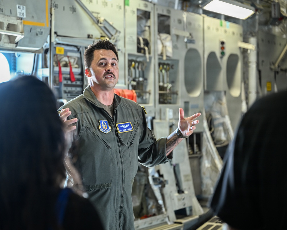 A U.S. Air Force airman in an olive green flight suit is speaking and gesturing to a small group of people inside what appears to be a military aircraft cargo bay. He is wearing unit patches and a name tag, suggesting he may be conducting a briefing or tour. The setting is the interior of a large military transport aircraft with visible equipment panels and systems.