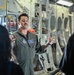 A U.S. Air Force airman in an olive green flight suit is speaking and gesturing to a small group of people inside what appears to be a military aircraft cargo bay. He is wearing unit patches and a name tag, suggesting he may be conducting a briefing or tour. The setting is the interior of a large military transport aircraft with visible equipment panels and systems.