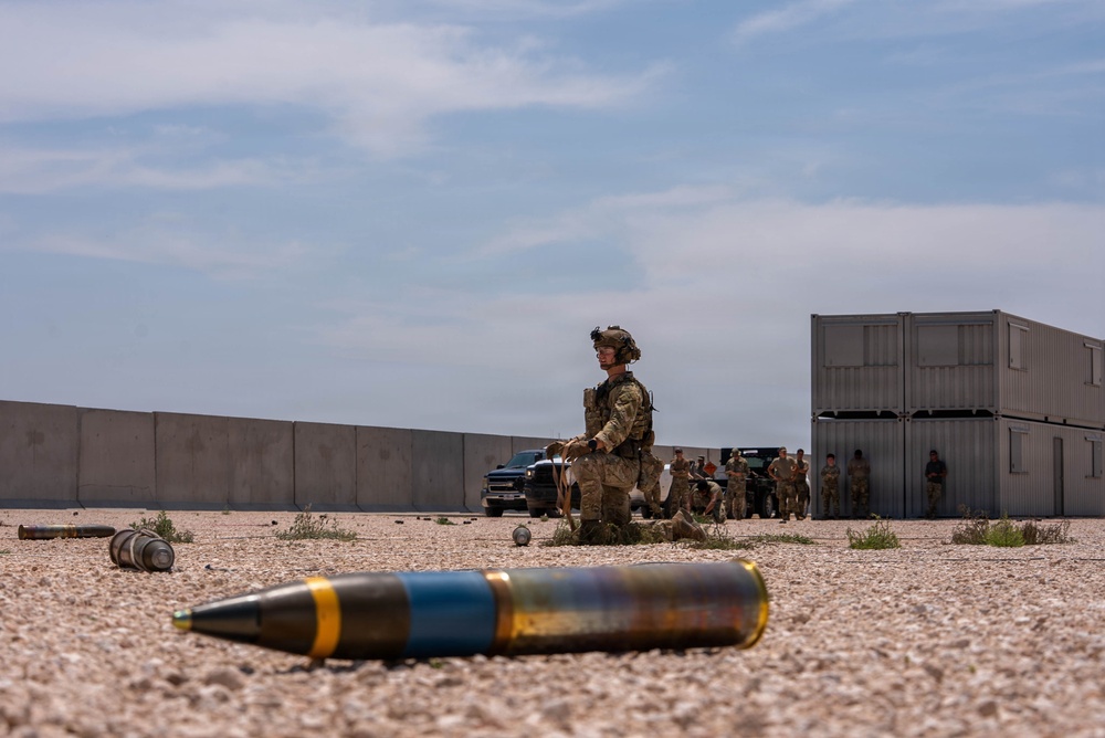 A military training or operational scene set in an arid environment, featuring a large-caliber projectile in the foreground with blue and gold markings. A soldier in full combat gear and helmet stands in the mid-ground, with additional personnel and military vehicles visible in the background. The setting includes concrete blast walls and stacked shipping containers consistent with a forward operating base or training range.