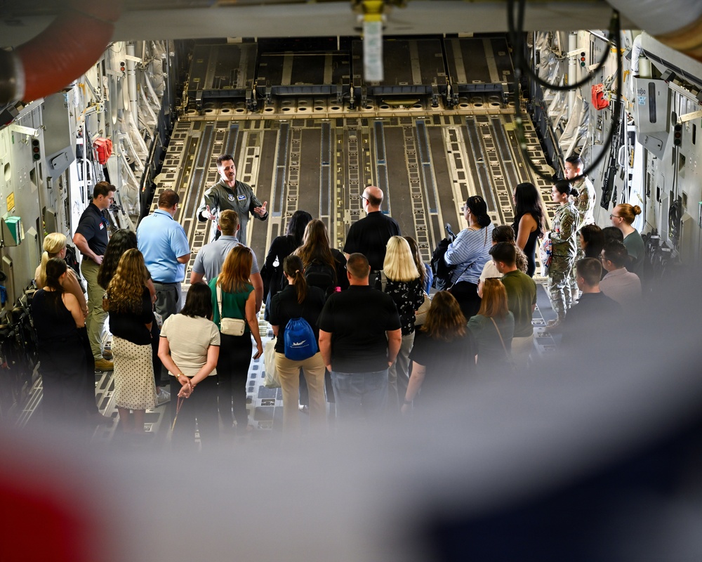 A military personnel member in flight suit is briefing a large group of civilians inside the cargo hold of a large military transport aircraft, likely a C-17 Globemaster III. The group of approximately 25-30 people are standing and listening to the presentation near the rear cargo ramp area. This appears to be an official tour or community outreach event at a military installation.