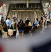 A military personnel member in flight suit is briefing a large group of civilians inside the cargo hold of a large military transport aircraft, likely a C-17 Globemaster III. The group of approximately 25-30 people are standing and listening to the presentation near the rear cargo ramp area. This appears to be an official tour or community outreach event at a military installation.
