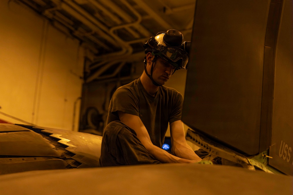 A military service member wearing a helmet with mounted goggles is working on what appears to be a naval aircraft in a ship's hangar bay, illuminated by warm amber lighting. The letters 'USS' are partially visible on a surface in the background, confirming a U.S. Navy vessel setting. The technician appears to be performing maintenance or inspection work on the aircraft's fuselage or wing surface.
