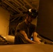A military service member wearing a helmet with mounted goggles is working on what appears to be a naval aircraft in a ship's hangar bay, illuminated by warm amber lighting. The letters 'USS' are partially visible on a surface in the background, confirming a U.S. Navy vessel setting. The technician appears to be performing maintenance or inspection work on the aircraft's fuselage or wing surface.