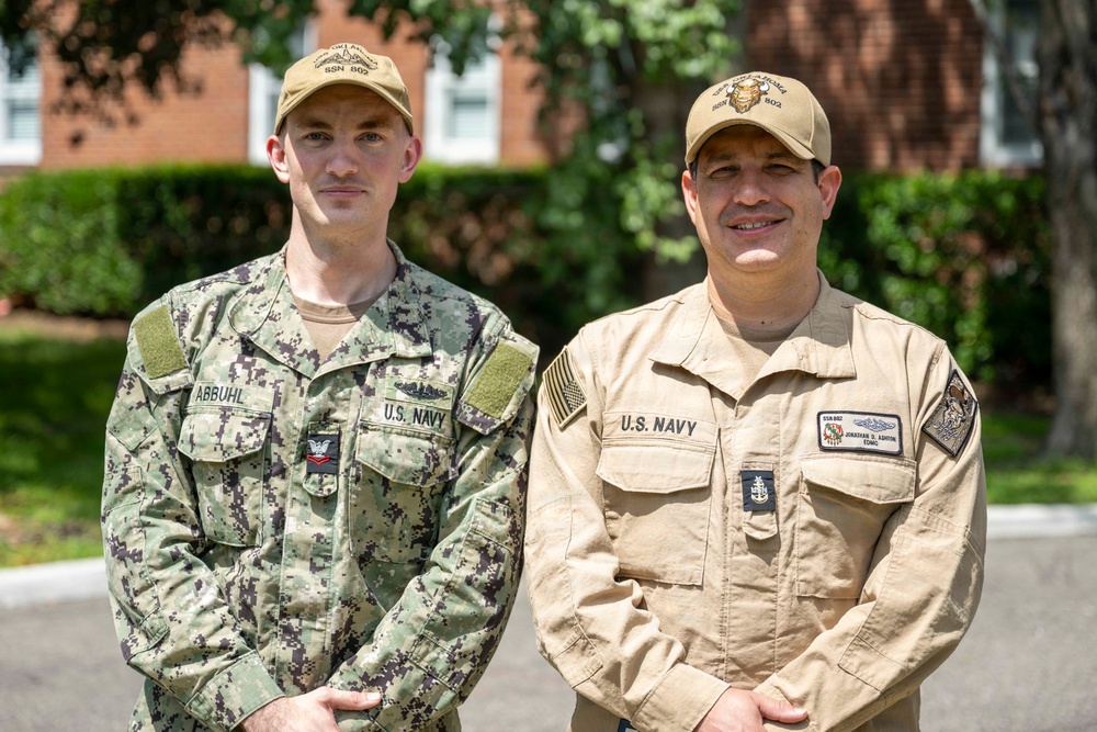 Two U.S. Navy personnel are posing together outdoors in front of a brick building with green foliage in the background. The sailor on the left wears the Navy Working Uniform (NWU) digital camouflage pattern, while the sailor on the right wears the tan/khaki Navy coverall uniform. Both wear matching caps with 'SSN 802' visible, indicating assignment to a submarine.