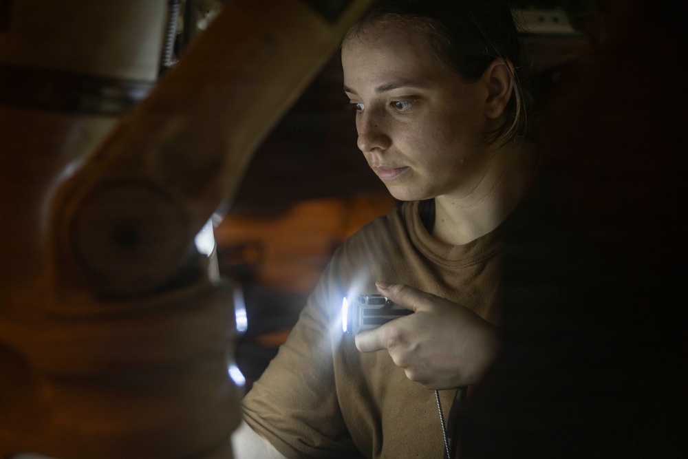 A young woman is examining an object in a dark environment using a small handheld flashlight or light source. The low-light conditions suggest she is inspecting something in a dimly lit space such as a storage area, attic, or garage. Her focused expression indicates careful, deliberate examination of the subject in front of her.