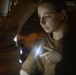A young woman is examining an object in a dark environment using a small handheld flashlight or light source. The low-light conditions suggest she is inspecting something in a dimly lit space such as a storage area, attic, or garage. Her focused expression indicates careful, deliberate examination of the subject in front of her.