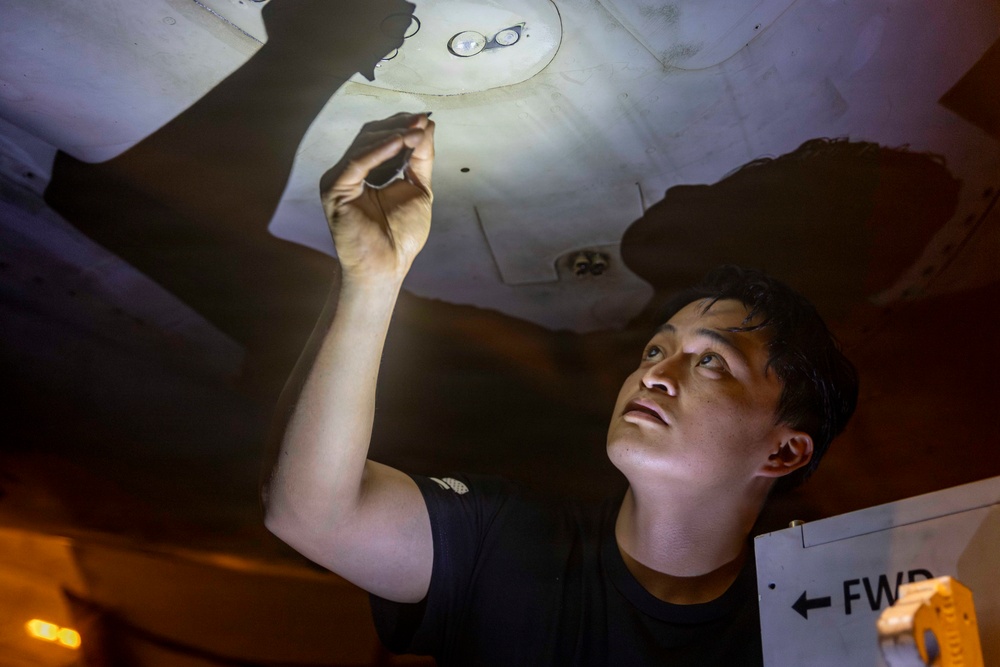A young man is working overhead, reaching up to inspect or service a component on the underside of a surface, likely an aircraft or vehicle. He is illuminated by a bright work light, casting dramatic shadows. A directional sign reading 'FWD' with an arrow is visible in the lower right corner, suggesting a military or aviation maintenance context.