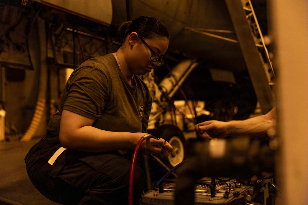 A female military technician or mechanic is performing maintenance work on what appears to be a military aircraft in a hangar or flight deck environment. She is working with colored hoses (red and blue) connected to a service unit or test equipment. The scene is lit with warm amber/orange lighting, suggesting an enclosed hangar bay, possibly aboard a naval vessel.