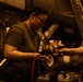A female military technician or mechanic is performing maintenance work on what appears to be a military aircraft in a hangar or flight deck environment. She is working with colored hoses (red and blue) connected to a service unit or test equipment. The scene is lit with warm amber/orange lighting, suggesting an enclosed hangar bay, possibly aboard a naval vessel.