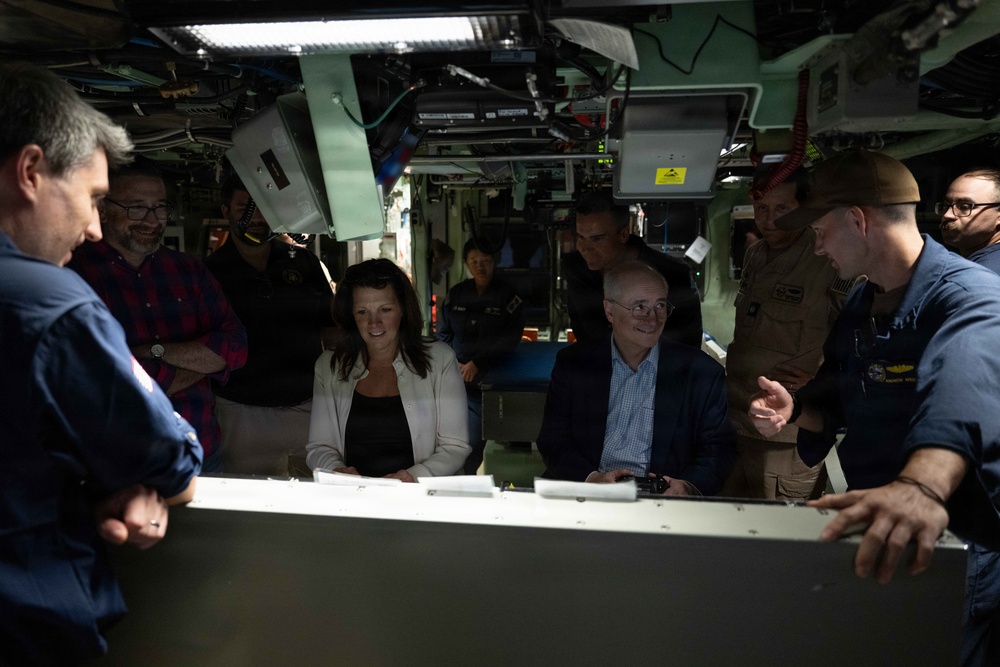 A group of civilians and military personnel are gathered inside what appears to be a submarine or naval vessel interior. They are engaged in a briefing or tour, gathered around a console or table in a cramped, equipment-filled space. The atmosphere appears informal and conversational, with several people smiling.