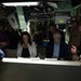 A group of civilians and military personnel are gathered inside what appears to be a submarine or naval vessel interior. They are engaged in a briefing or tour, gathered around a console or table in a cramped, equipment-filled space. The atmosphere appears informal and conversational, with several people smiling.