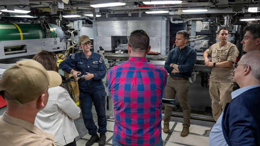 A U.S. Navy sailor in uniform is briefing a group of civilians and at least one other uniformed Navy personnel inside what appears to be a naval vessel's interior compartment. The setting contains large green munitions or ordnance visible in the background, suggesting this is a weapons or ordnance storage area. The group appears to be on an official tour or familiarization visit.