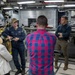 A U.S. Navy sailor in uniform is briefing a group of civilians and at least one other uniformed Navy personnel inside what appears to be a naval vessel's interior compartment. The setting contains large green munitions or ordnance visible in the background, suggesting this is a weapons or ordnance storage area. The group appears to be on an official tour or familiarization visit.