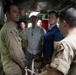 A group of approximately six individuals are gathered inside what appears to be a military galley or food service area aboard a naval vessel or military facility. The group includes personnel in military uniforms (tan/khaki Navy or similar branch uniforms and one Marine Corps camouflage uniform) alongside civilians in casual attire. One uniformed individual appears to be briefing or presenting information to the group.