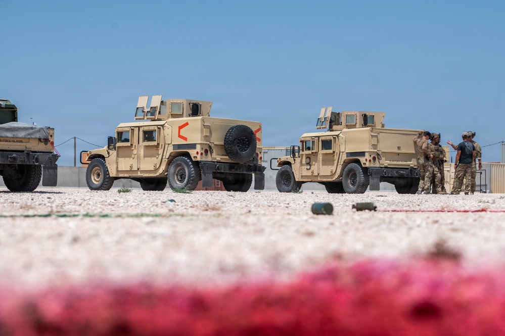 Two desert-tan military HMMWVs (Humvees) are parked on a gravel surface in what appears to be a forward operating base or military installation in an arid environment. The vehicles display red chevron/arrow markings on their rear doors, and a small group of personnel in military and civilian attire are gathered near the second vehicle. The foreground shows blurred red and white objects at ground level, likely rope or barrier material.