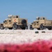 Two desert-tan military HMMWVs (Humvees) are parked on a gravel surface in what appears to be a forward operating base or military installation in an arid environment. The vehicles display red chevron/arrow markings on their rear doors, and a small group of personnel in military and civilian attire are gathered near the second vehicle. The foreground shows blurred red and white objects at ground level, likely rope or barrier material.