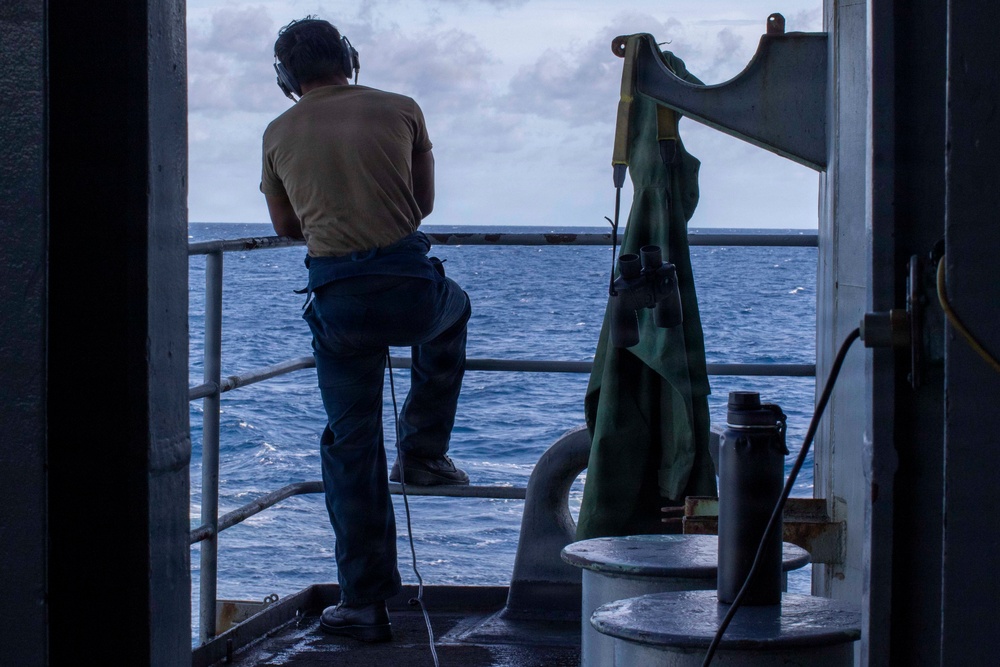 A person wearing a tan t-shirt, dark trousers, and communication headset stands on the exterior deck of a naval or military vessel, looking out over open ocean. The scene is photographed from inside a doorway or hatch, creating a silhouette effect against the bright sea and cloudy sky. Various maritime equipment including binoculars, a green cover or bag, and a thermos are visible nearby.