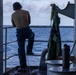 A person wearing a tan t-shirt, dark trousers, and communication headset stands on the exterior deck of a naval or military vessel, looking out over open ocean. The scene is photographed from inside a doorway or hatch, creating a silhouette effect against the bright sea and cloudy sky. Various maritime equipment including binoculars, a green cover or bag, and a thermos are visible nearby.