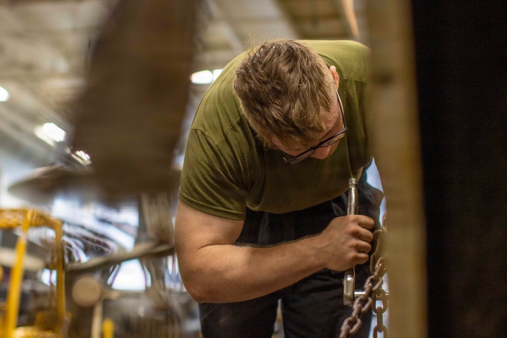 A man wearing a green t-shirt and glasses is bent over, working with a heavy chain and metal hardware in what appears to be an industrial or hangar setting. He is applying significant physical effort to manipulate the chain assembly. The background shows blurred industrial equipment and overhead lighting consistent with a large workshop or aircraft hangar.