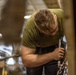 A man wearing a green t-shirt and glasses is bent over, working with a heavy chain and metal hardware in what appears to be an industrial or hangar setting. He is applying significant physical effort to manipulate the chain assembly. The background shows blurred industrial equipment and overhead lighting consistent with a large workshop or aircraft hangar.