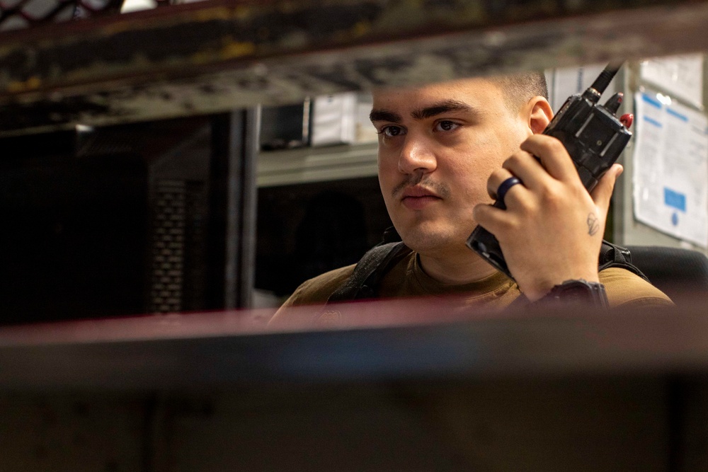 A young man in a tan/olive military-style shirt is holding a tactical handheld radio (walkie-talkie) up to his ear, appearing to be actively listening or communicating. The image is captured through a narrow gap between shelves or equipment racks, creating a framed, close-up perspective. The background suggests an industrial or military operational environment.