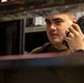 A young man in a tan/olive military-style shirt is holding a tactical handheld radio (walkie-talkie) up to his ear, appearing to be actively listening or communicating. The image is captured through a narrow gap between shelves or equipment racks, creating a framed, close-up perspective. The background suggests an industrial or military operational environment.