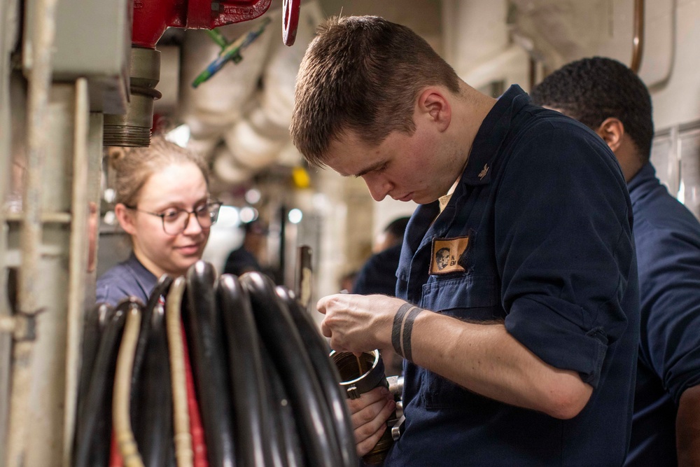 The image shows what appears to be U.S. Navy personnel in a shipboard compartment performing maintenance or technical work. A male sailor in the foreground is focused on manipulating a metal component, while a female sailor wearing glasses observes closely. The setting appears to be a narrow passageway or workspace aboard a naval vessel, identifiable by the pipes, cables, and confined industrial environment.