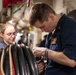 The image shows what appears to be U.S. Navy personnel in a shipboard compartment performing maintenance or technical work. A male sailor in the foreground is focused on manipulating a metal component, while a female sailor wearing glasses observes closely. The setting appears to be a narrow passageway or workspace aboard a naval vessel, identifiable by the pipes, cables, and confined industrial environment.