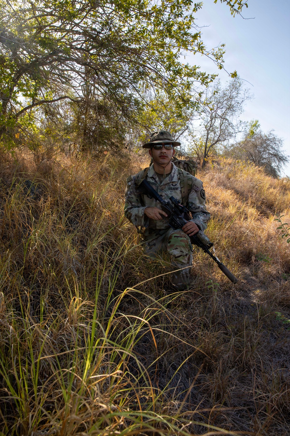 A U.S. Army soldier in full Operational Camouflage Pattern (OCP) uniform is kneeling in a dry, brushy terrain while holding an assault rifle. The soldier is wearing a boonie hat, sunglasses, and a rucksack, suggesting a field training exercise or patrol operation. The environment appears to be arid scrubland with dry grass and sparse trees under a clear sky.